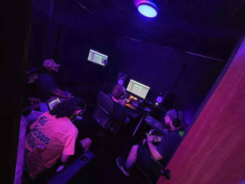 Everybody on something — laptop, phones, the desk — under the purple ceiling light